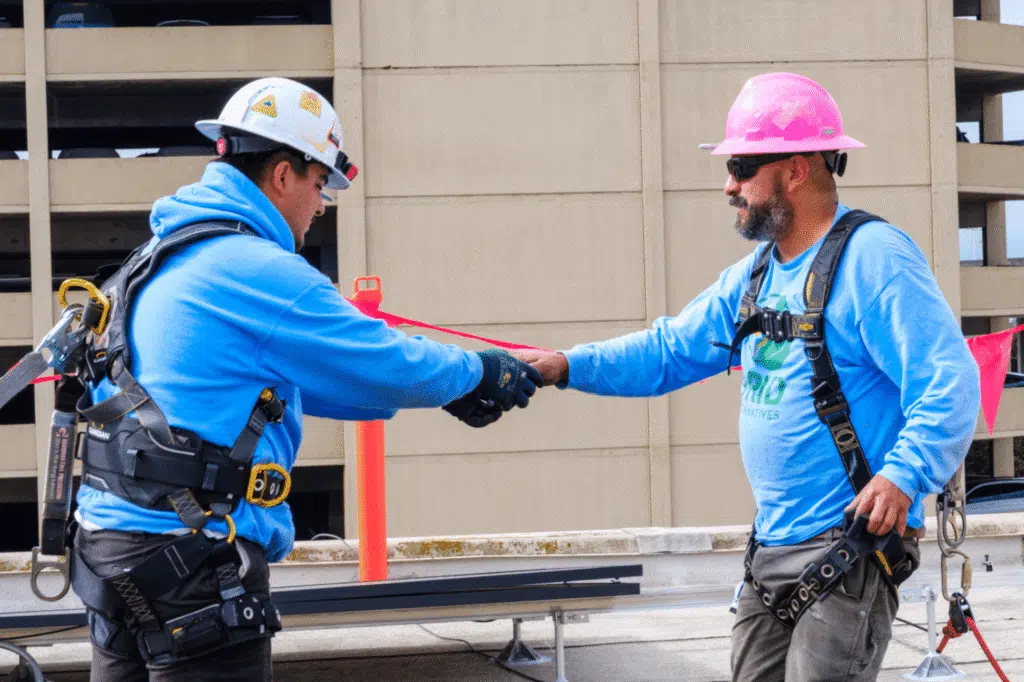 Two installers in hard hats passing a red strap between them on a rooftop, a parking structure behind.