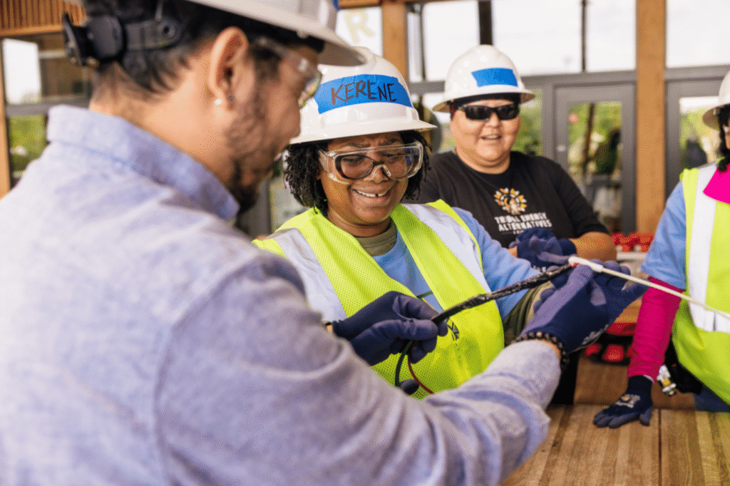 An instructor in a hard hat showing a trainee in a blue hard hat how to handle a tool, others watching.