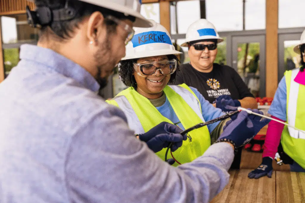 An instructor in a hard hat showing a trainee in a blue hard hat how to handle a tool, others watching.