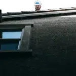 A worker in a white hard hat looks out over the ridge of a house roof, seen from the ground below.