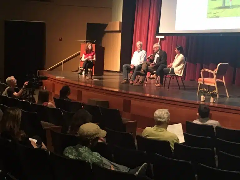 Panel discussion on stage with four speakers seated in chairs and audience members in foreground, one operating video camera