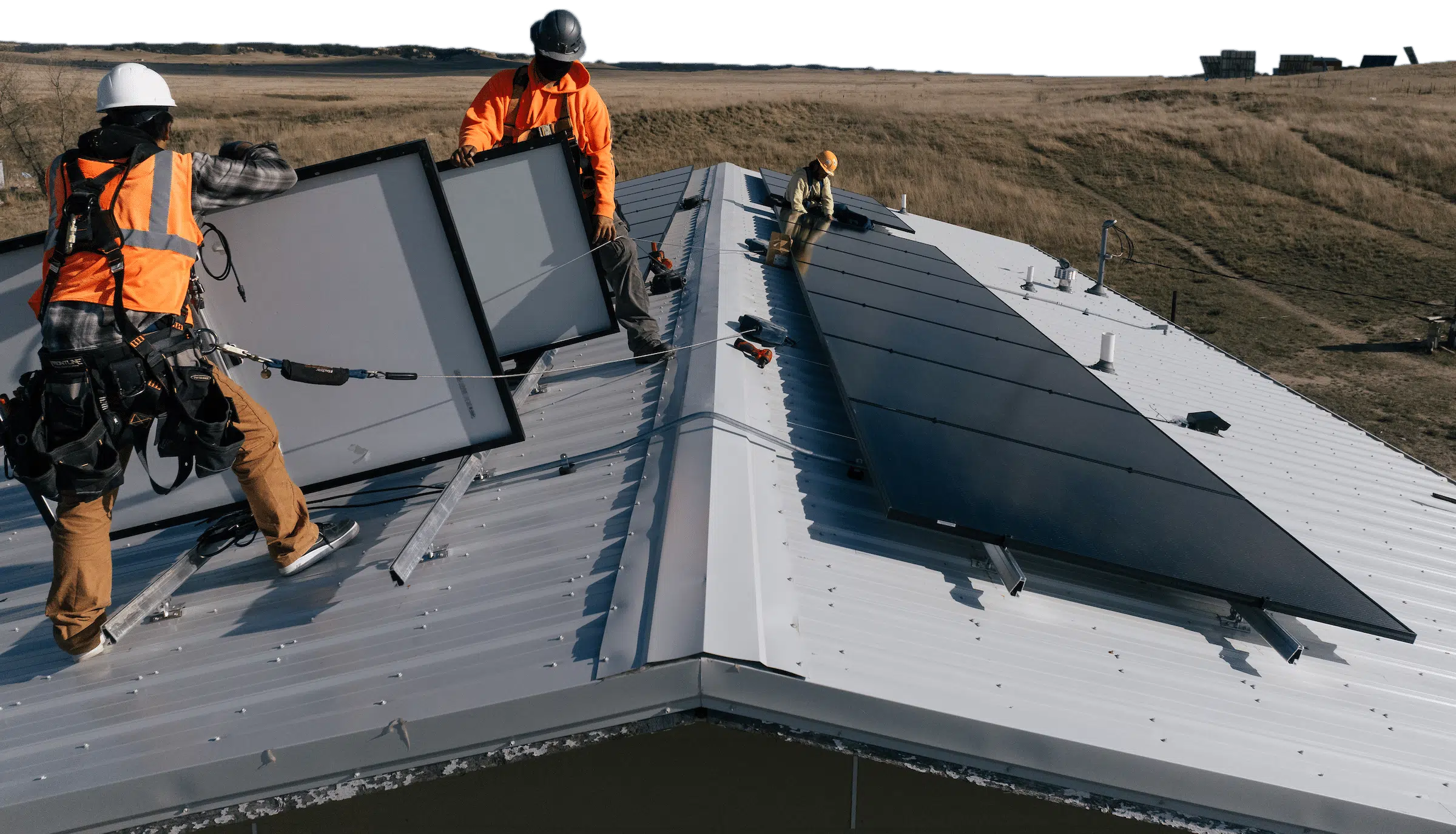 Three installers in high-visibility gear lifting and setting solar panels along the ridge of a metal barn roof.