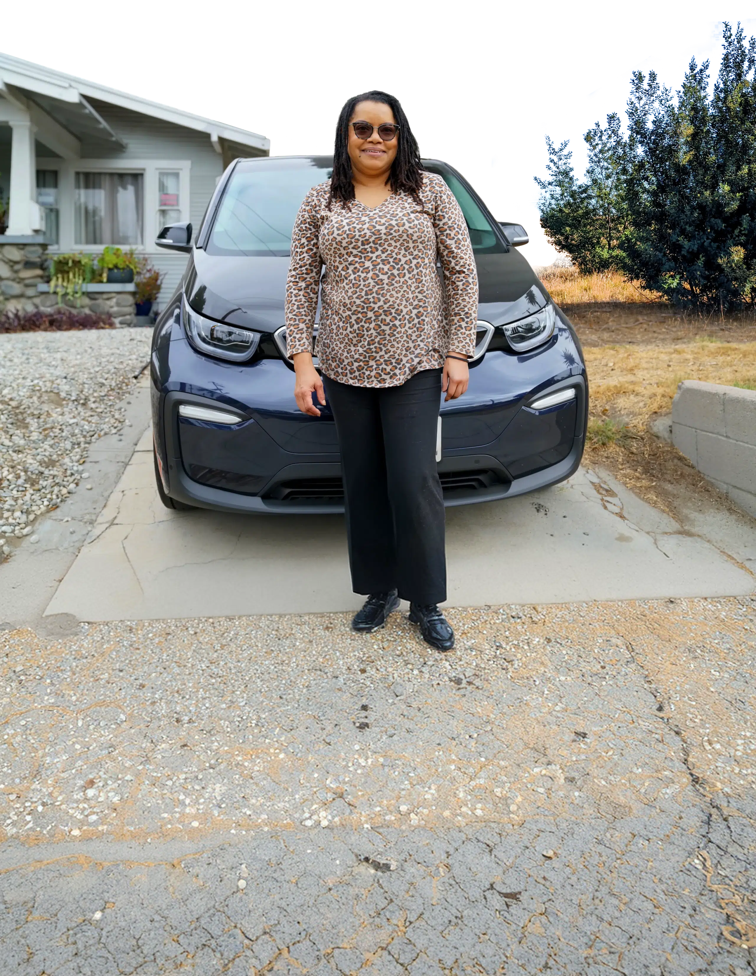 A woman standing beside her electric car in the driveway of a single-storey house.