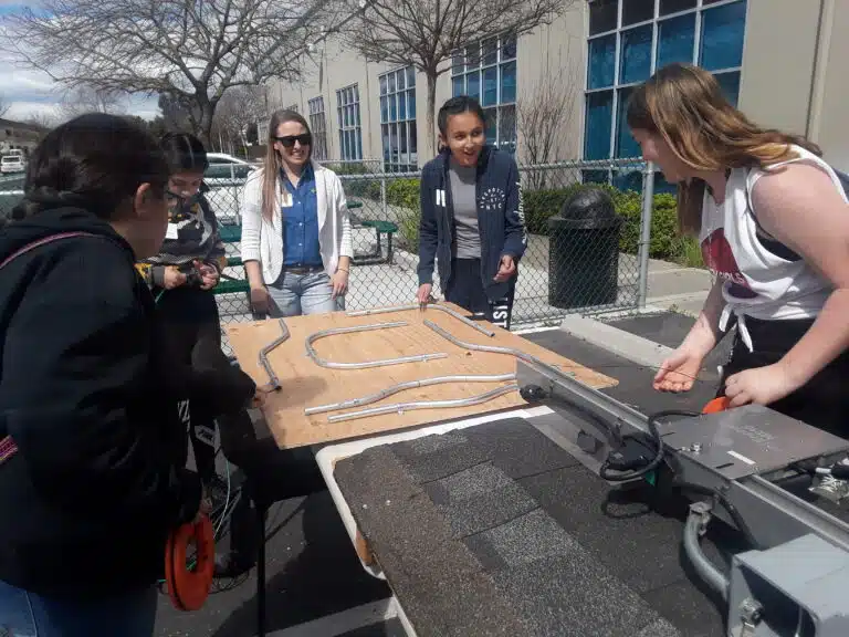 Five students examine a wooden circuit board model outdoors in a courtyard setting