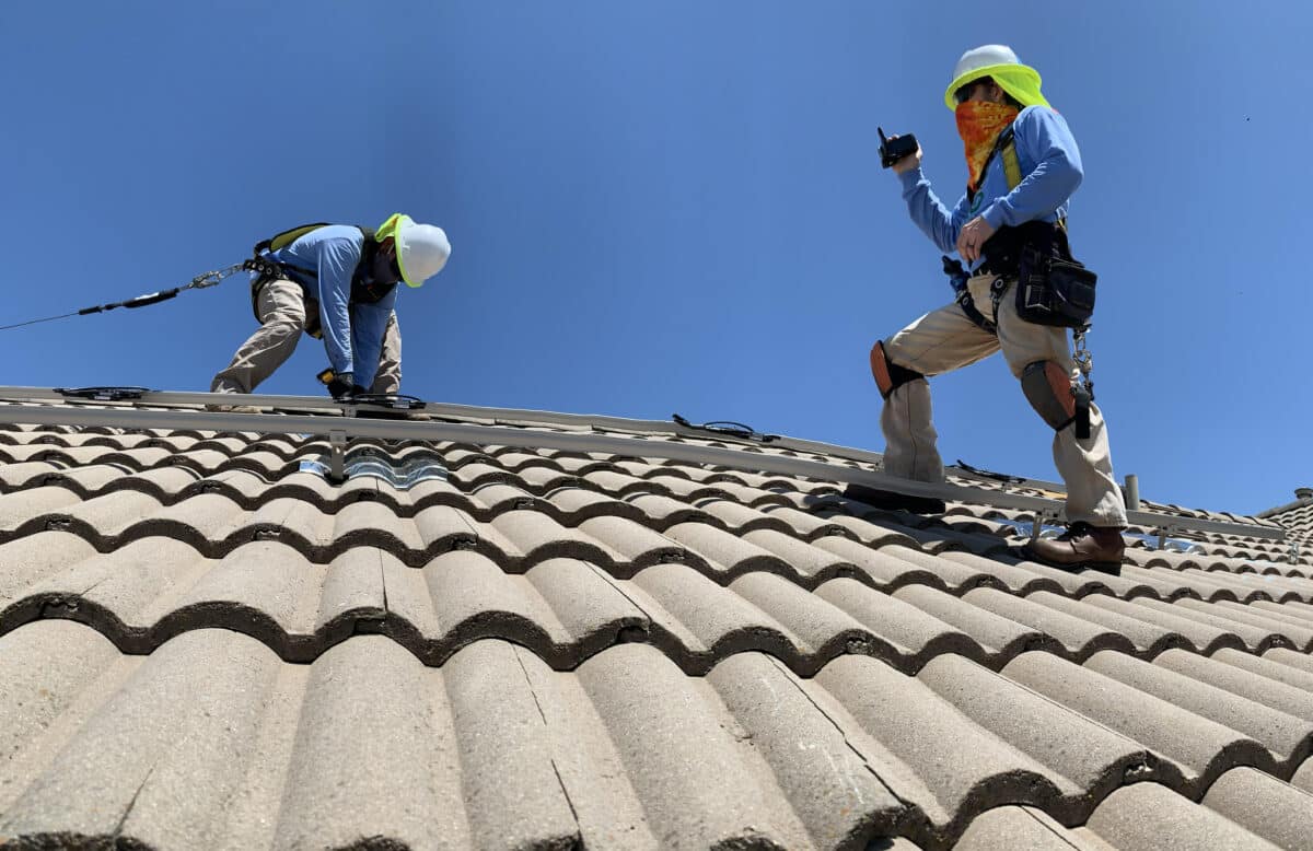 Two workers with safety gear performing roof tile installation on a steep residential roof under clear sky