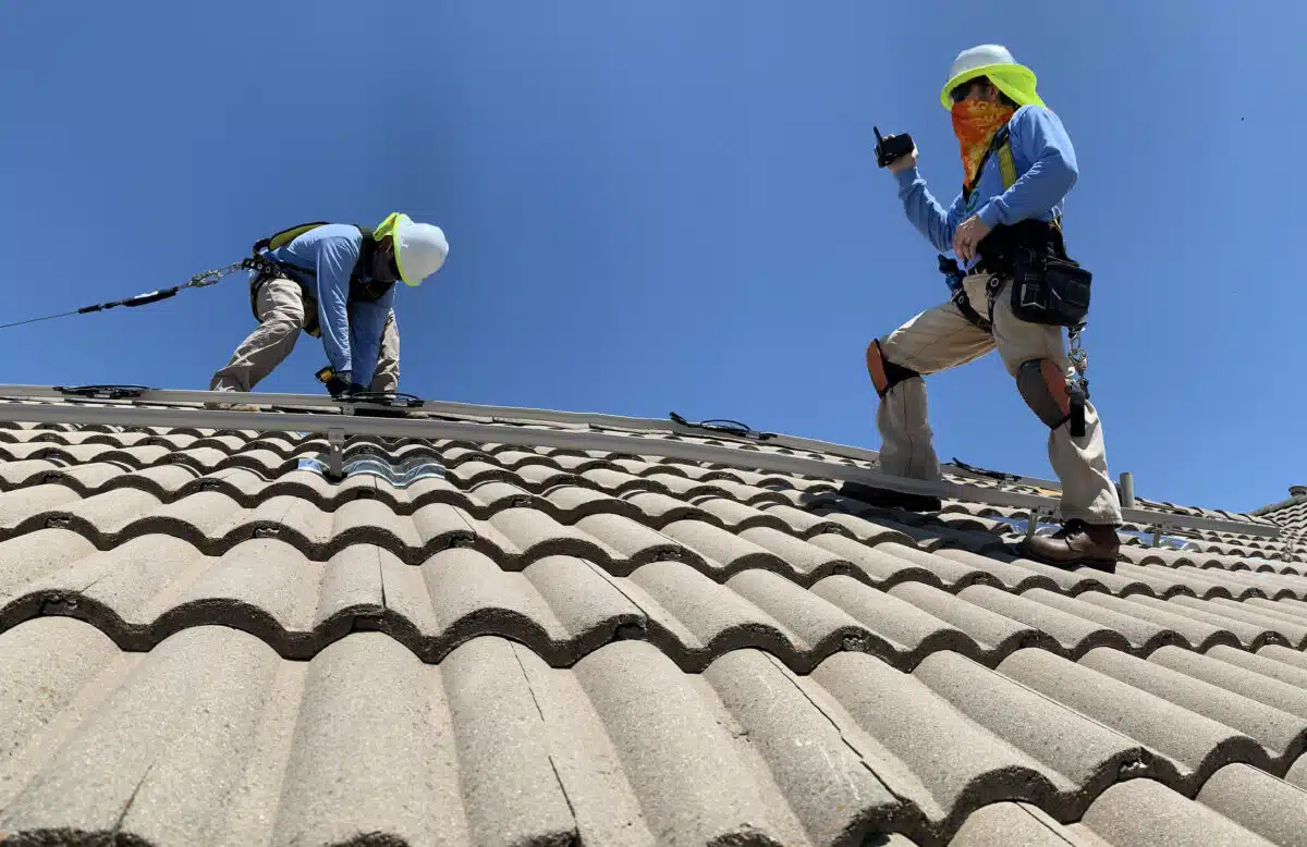 Two workers with safety gear performing roof tile installation on a steep residential roof under clear sky