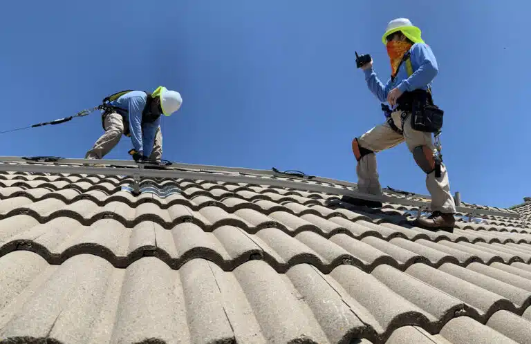Two workers with safety gear performing roof tile installation on a steep residential roof under clear sky
