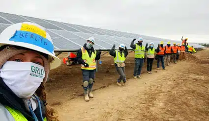 Worker in hard hat and mask at solar farm construction site with team in background