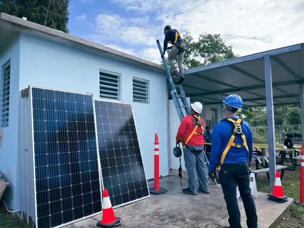 Installers in hard hats carry a solar panel past a carport frame on site.