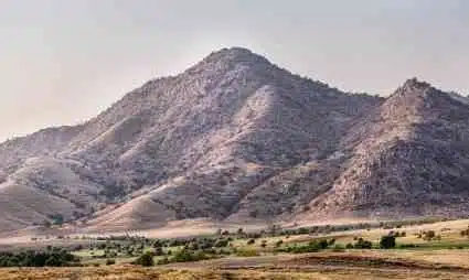 Rocky mountain peak with green valley and scattered trees below under cloudy sky