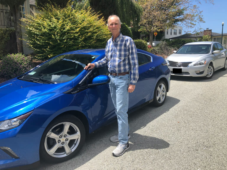 Man standing beside blue car in driveway with residential homes in background