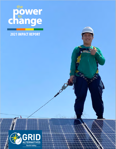 GRID NV Staff Angelo stands above some solar panels and smiles and gives the camera a thumbs up. He is wearing a green shirth and blue pants, with protective fall and head gear.
