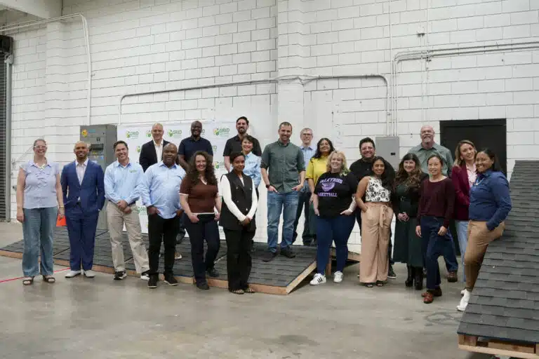 Group of approximately 25 people posing together in an industrial warehouse space with white brick walls and exposed pipes