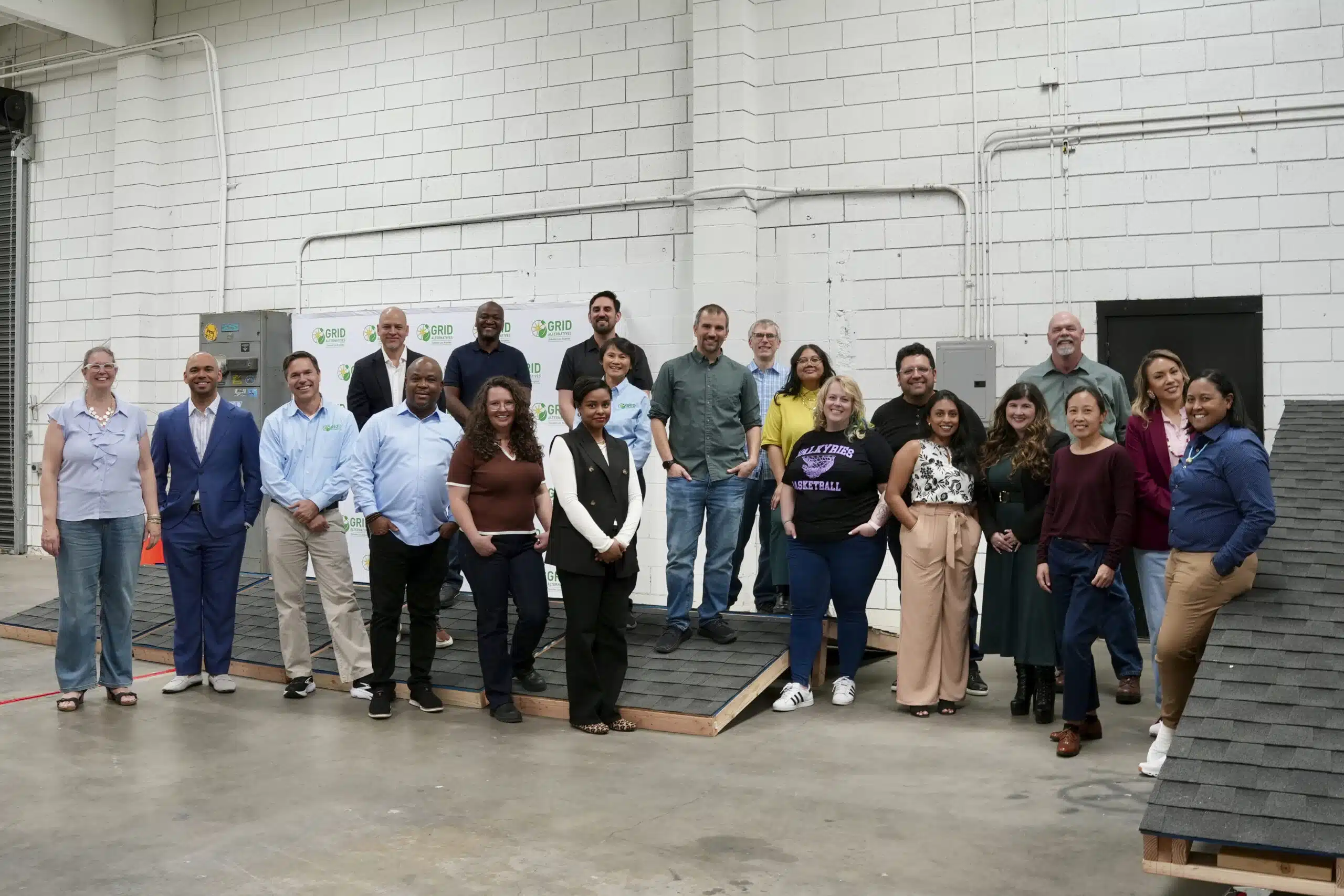 Group of approximately 25 people posing together in an industrial warehouse space with white brick walls and exposed pipes