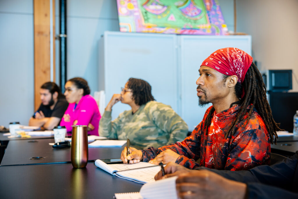 Man in red bandana and patterned shirt takes notes during classroom training session with four other participants seated at table