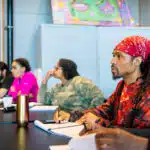 Man in red bandana and patterned shirt takes notes during classroom training session with four other participants seated at table