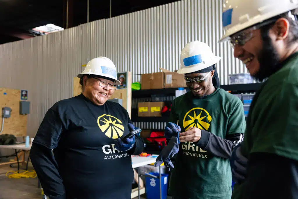 Three workers in hard hats and GRID Alternatives shirts smile together in a warehouse facility during training