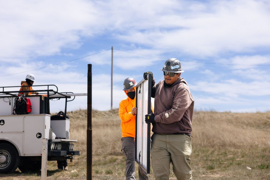 Workers in hard hats installing utility poles in a prairie landscape near a service truck