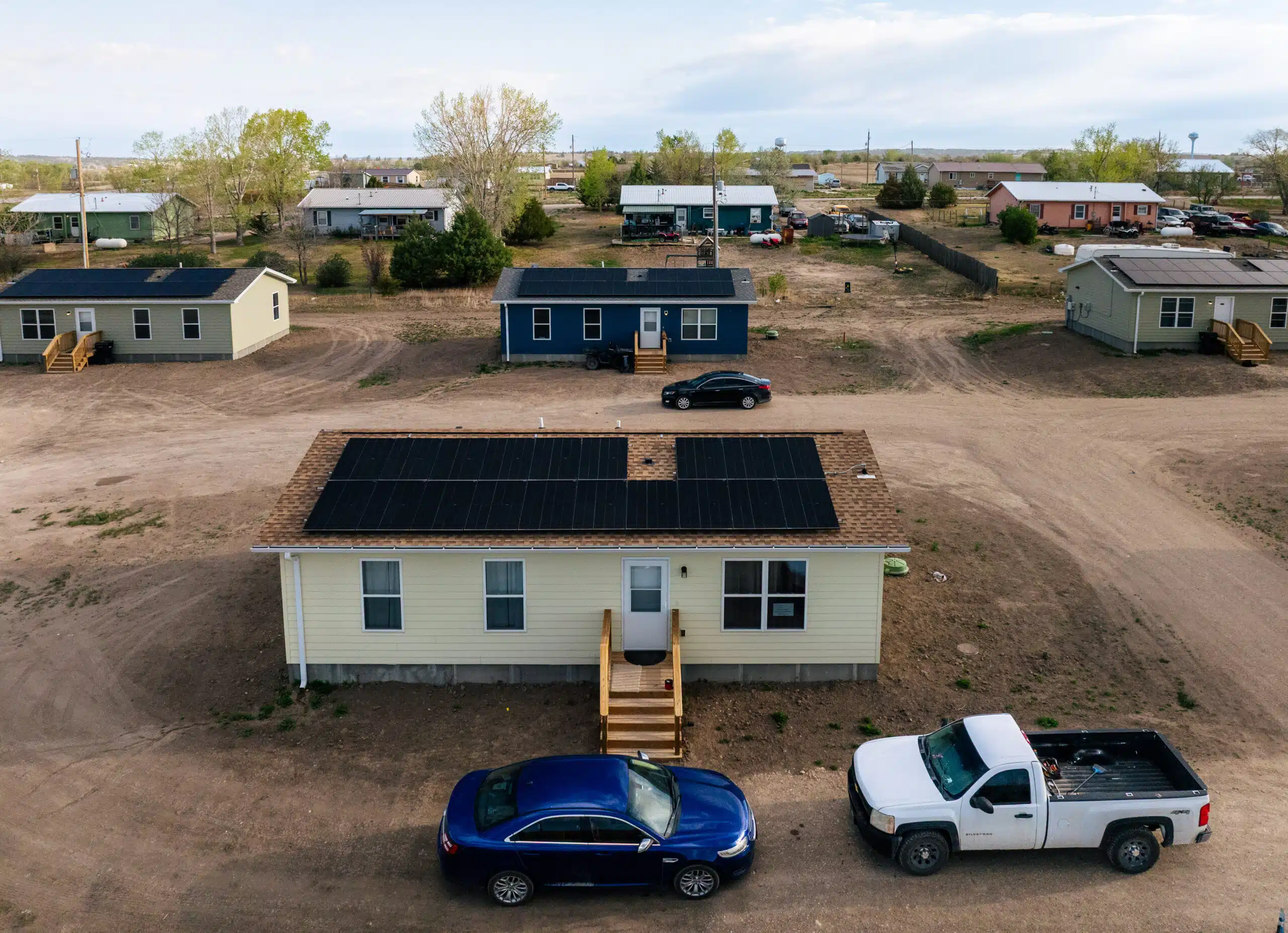 Cream-colored mobile home with solar panels on roof, parked cars in foreground, rural development with similar homes visible in background
