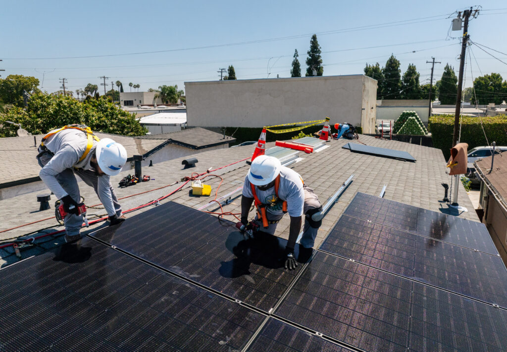 Two workers in white hard hats installing solar panels on a residential rooftop with safety equipment and ropes visible