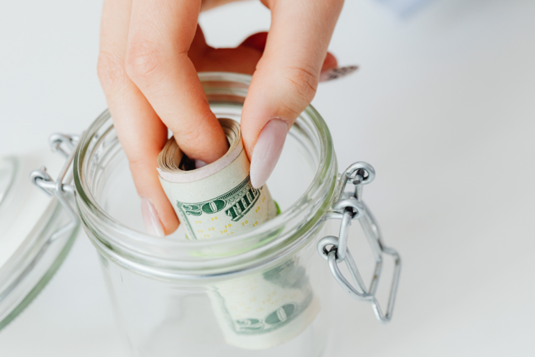 Hand placing a rolled $20 bill into a glass jar labeled with dollar signs