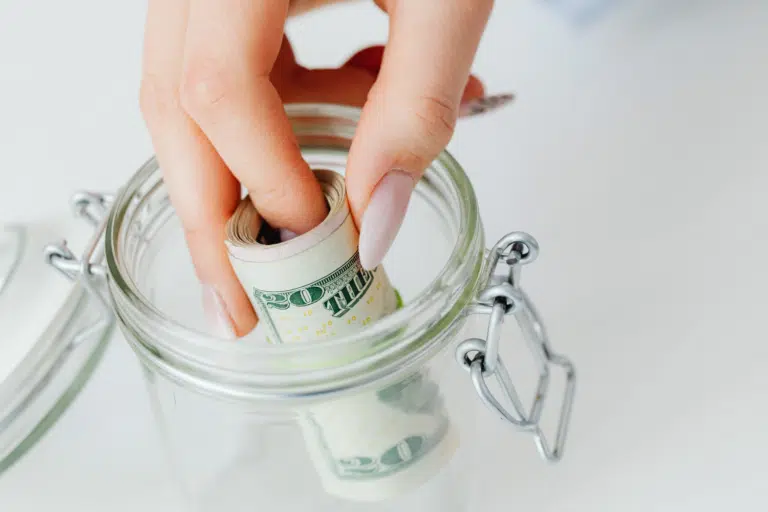 Hand placing a rolled $20 bill into a glass jar labeled with dollar signs