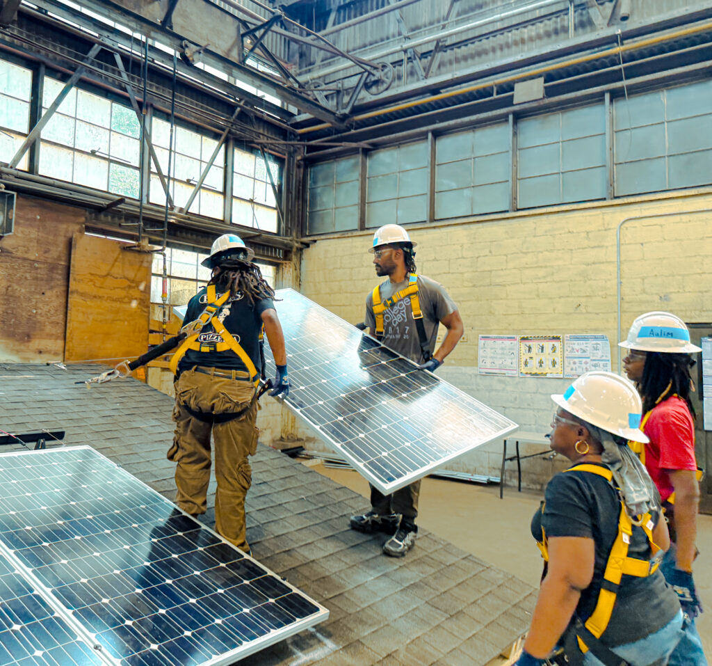 Four workers in hard hats and safety gear examining solar panels in an industrial warehouse training facility