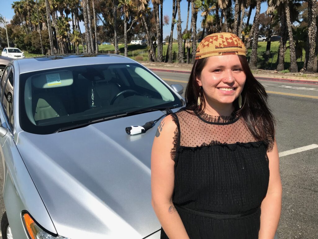 Smiling woman wearing orange headband stands next to white sedan in sunny parking lot lined with palm trees