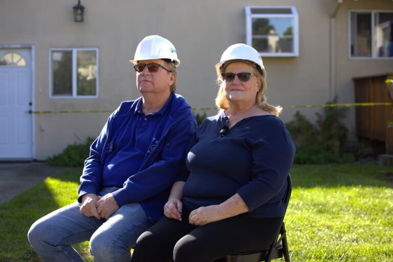 Two homeowner clients sitting in front of their home, wearing hard hats