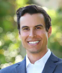Professional headshot of smiling man in navy blazer and white shirt against blurred green background