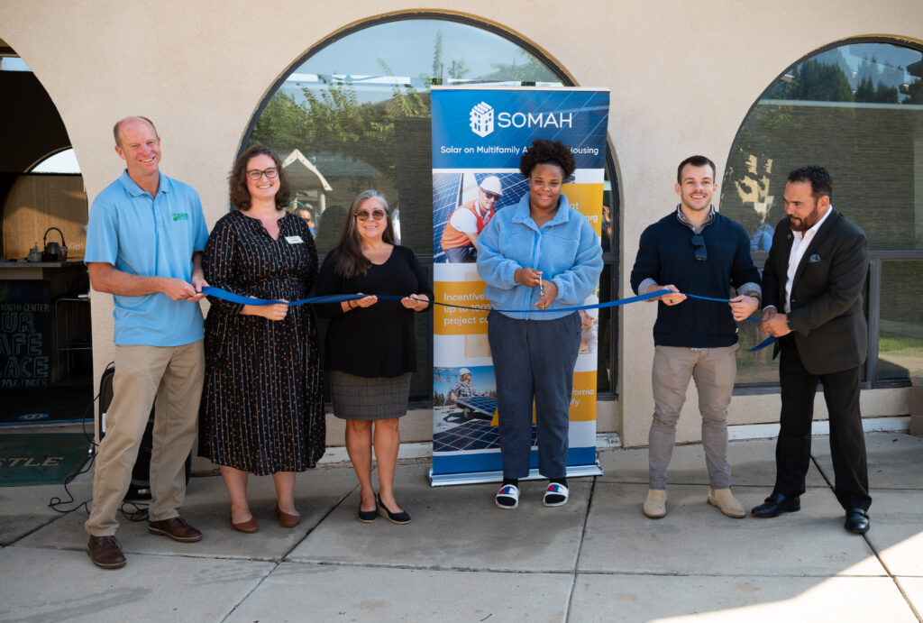 Six people cutting a blue ribbon at a SOMAH solar housing project ribbon-cutting ceremony