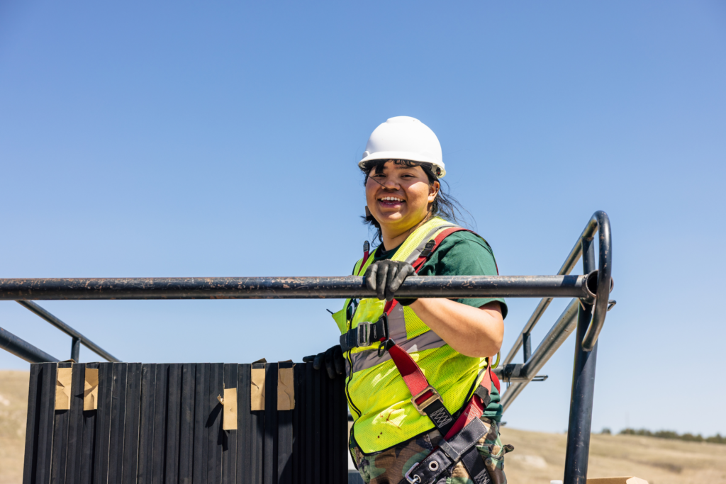 A GRID crew member in a hard hat and hi-vis vest smiles while leaning on the frame of a ground-mounted solar array