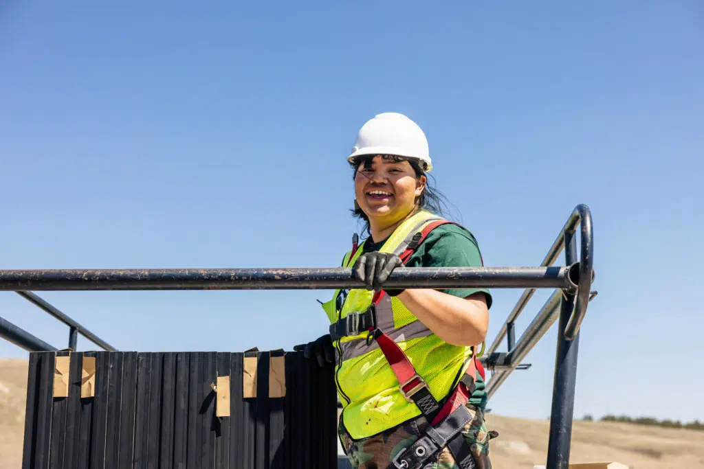 A GRID crew member in a hard hat and hi-vis vest smiles while leaning on the frame of a ground-mounted solar array