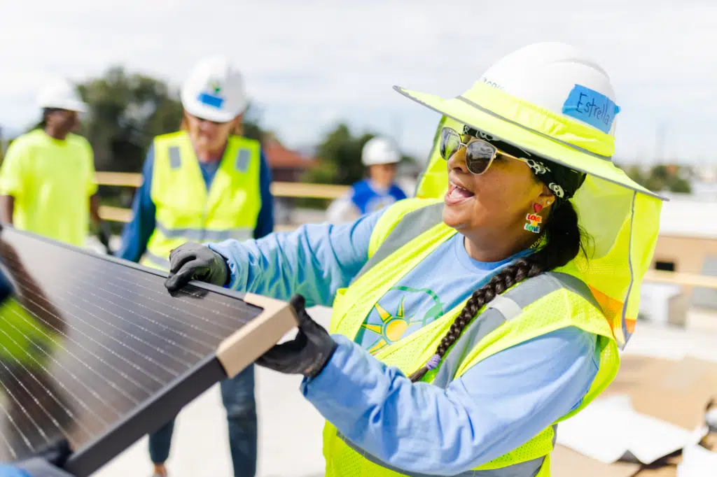 A GRID volunteer in a hard hat and hi-vis vest laughs as she guides a solar module into place, with three teammates working behind her