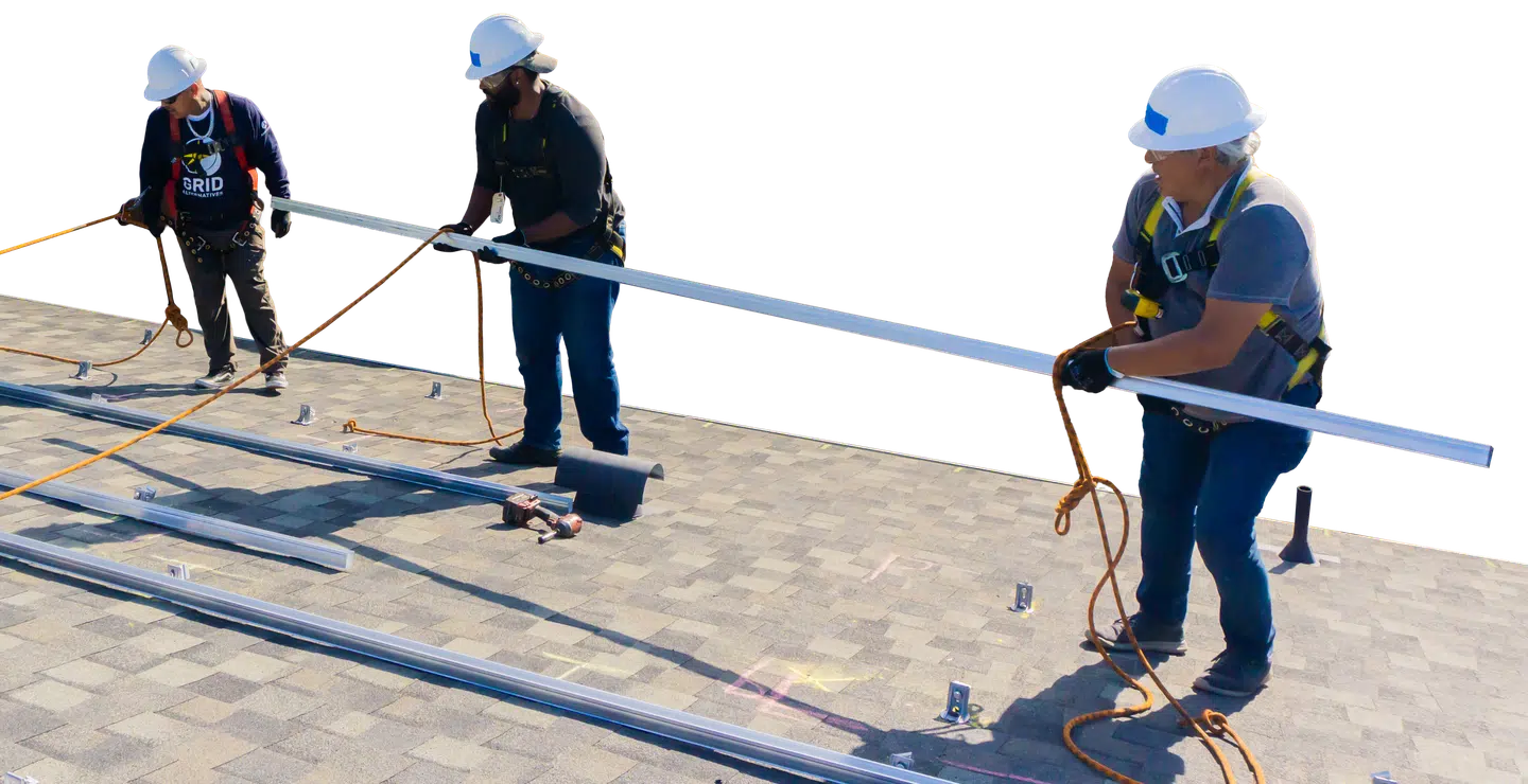 Three installers in hard hats and harnesses passing a length of racking rail across a shingle roof.