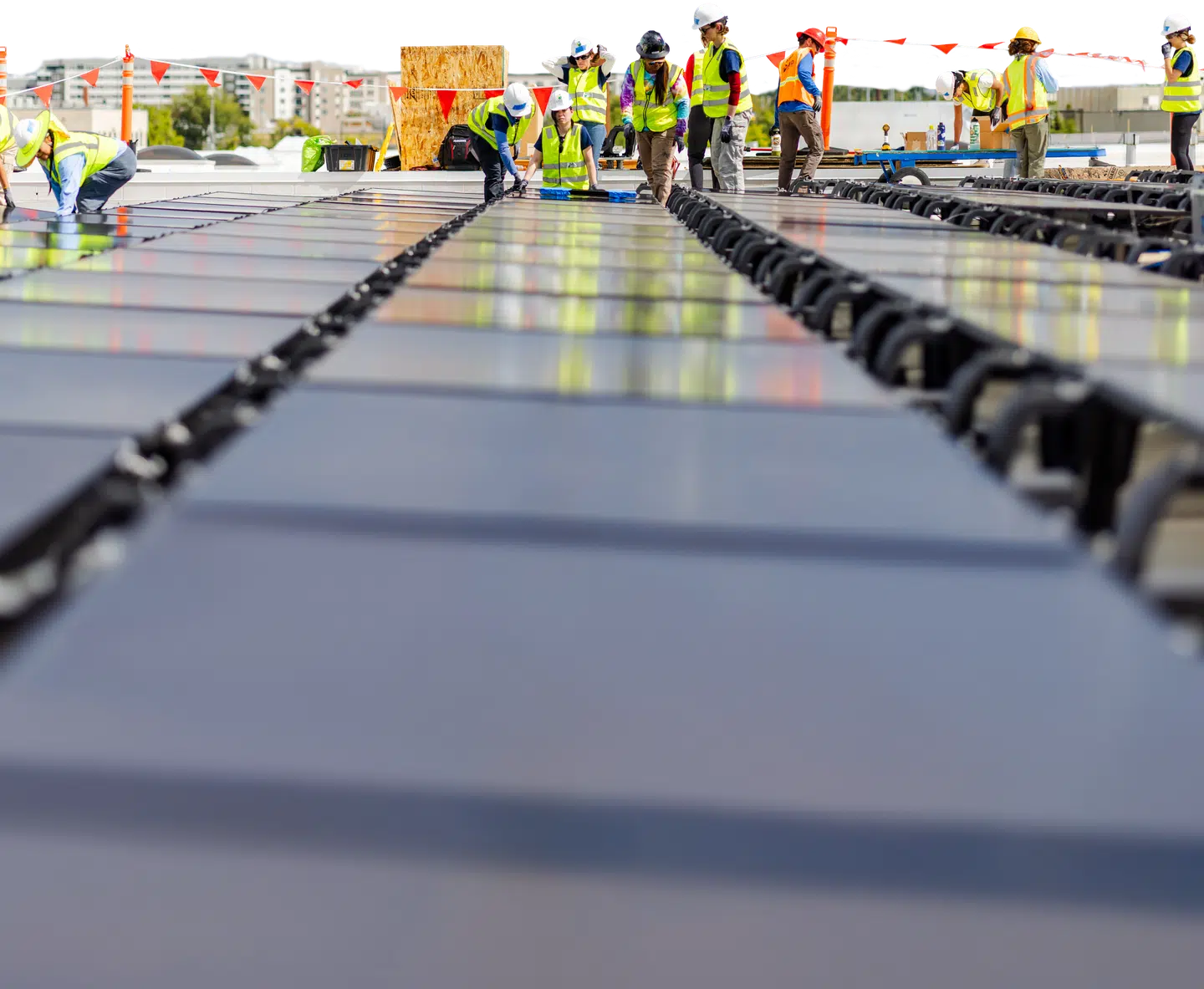 A crew of installers in hi-vis vests and hard hats working across rows of solar panels on a flat commercial roof, city skyline behind them.