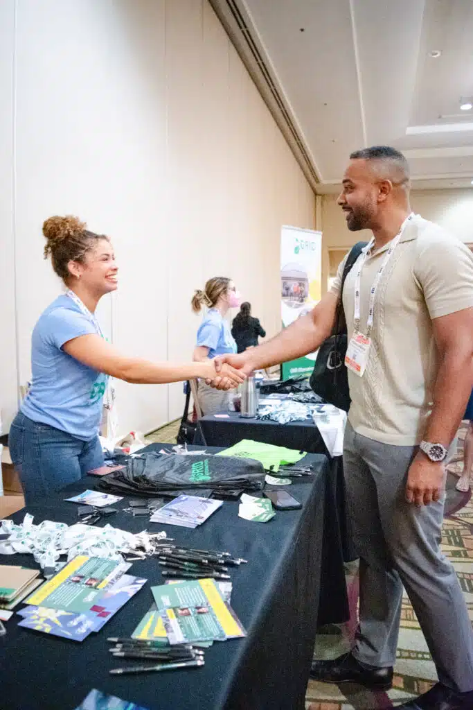 Two people shaking hands at a conference booth displaying promotional materials and merchandise on a black table