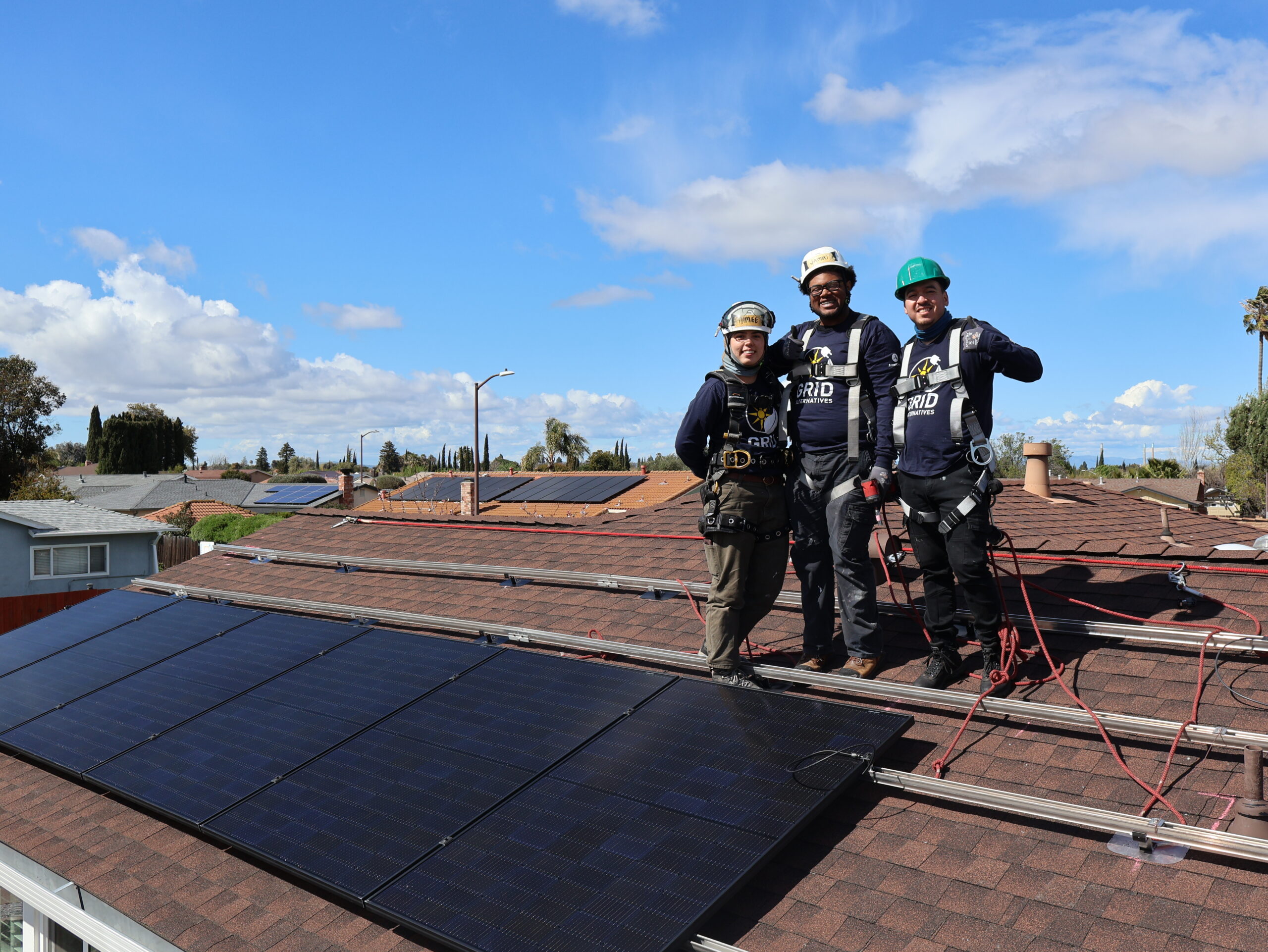 Three solar installation workers in safety gear stand on a residential rooftop with solar panels and blue sky