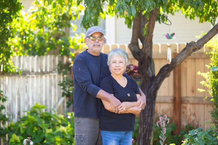 Older couple posing together in a sunny backyard garden with trees and wooden fence behind them