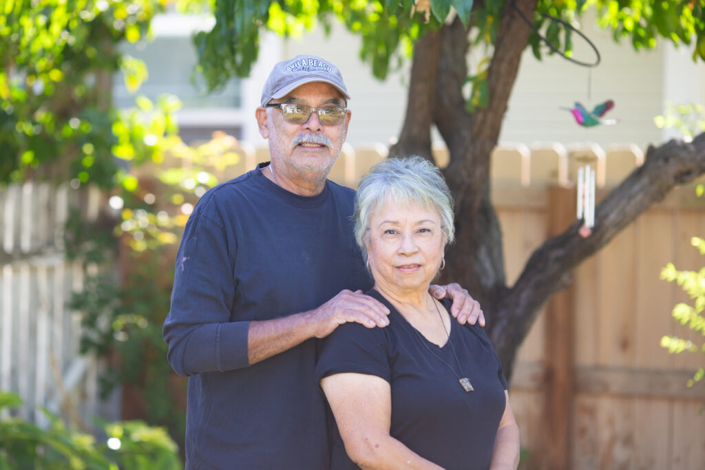 Older couple posing together under a leafy tree in a sunny backyard setting
