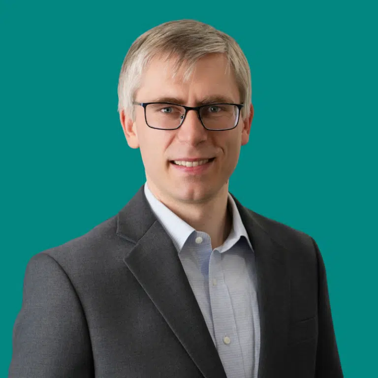 A man in a great blazer, with a light blue shirt, glasses, and blonde hair. A headshot against a deep teal background.