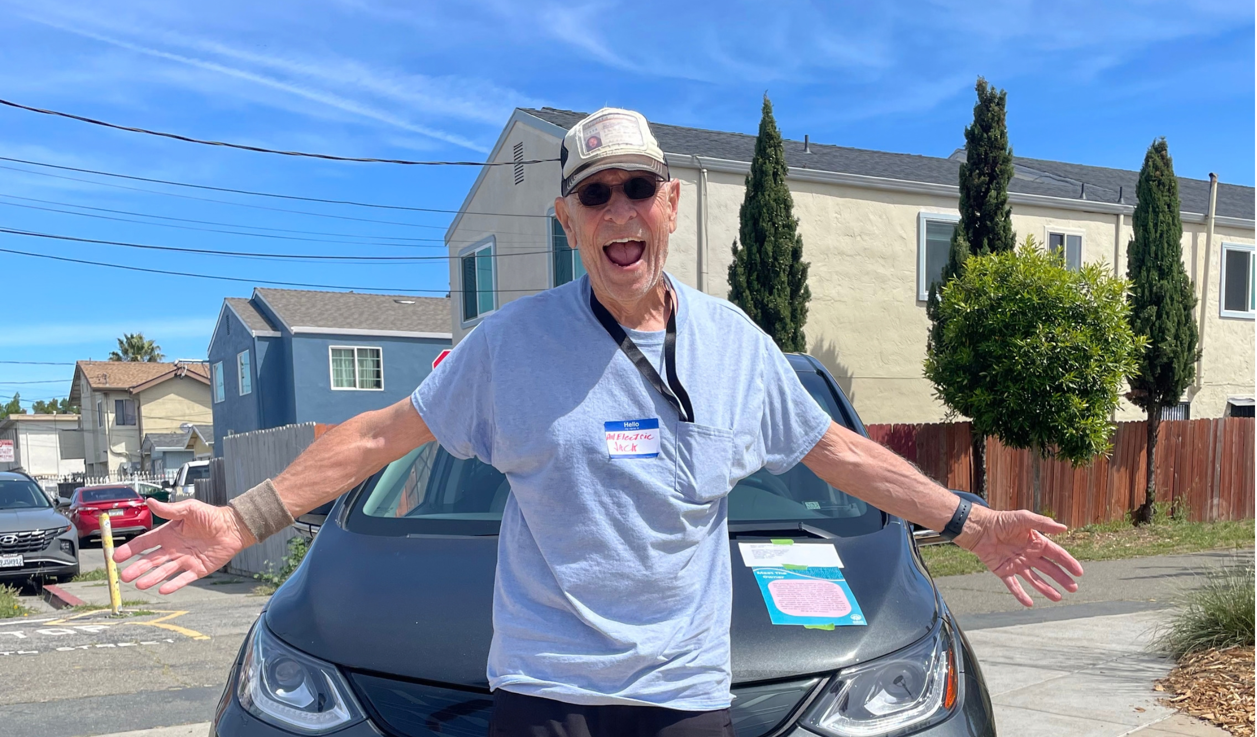 Man in cap and sunglasses standing in front of electric vehicle with arms spread wide, smiling at camera in residential neighborhood